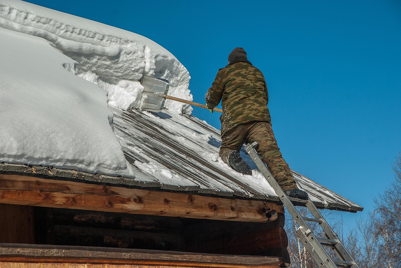 A worker on a ladder shoveling heavy snow off a wooden cabin roof